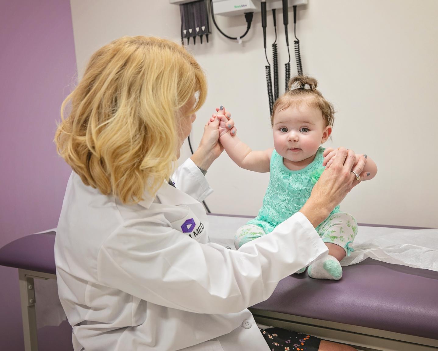 Infant sitting on an examination bed while holding a doctors hadns