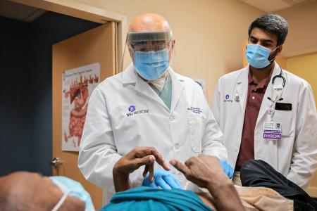 Two doctors wearing personal protective equipment examine a person who is laying on an examination table